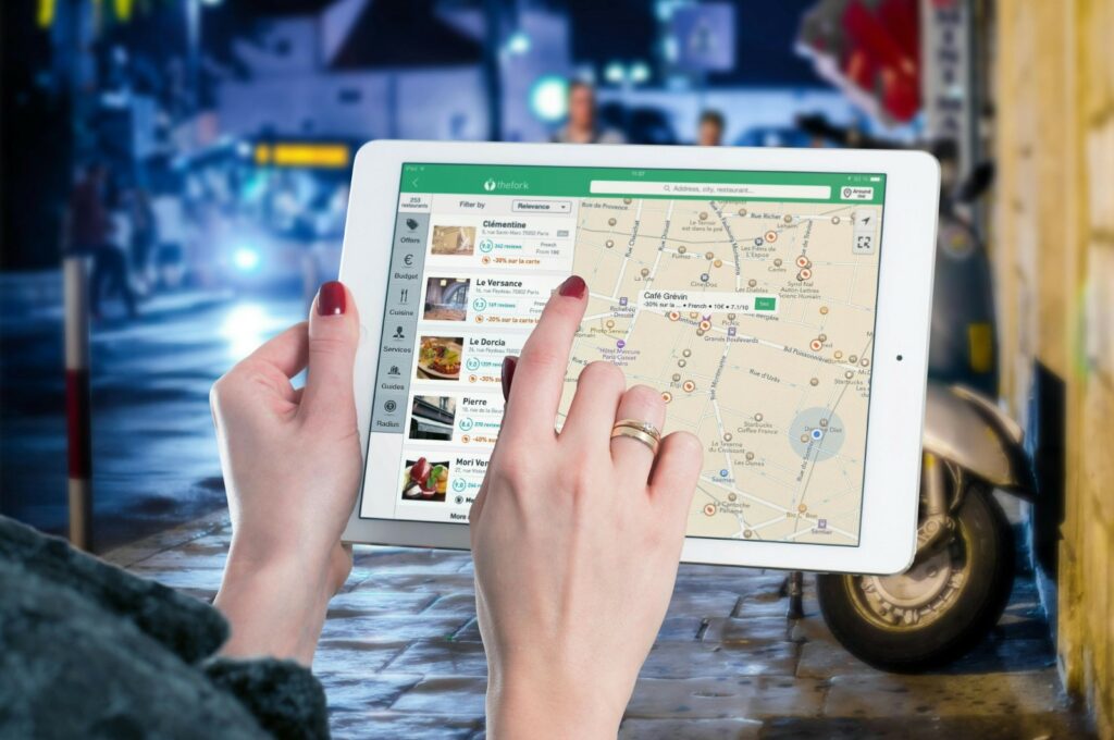 Close-up of a woman's hands holding a tablet displaying a map application, with potential graduation party venues highlighted, set against a lively street scene at night.