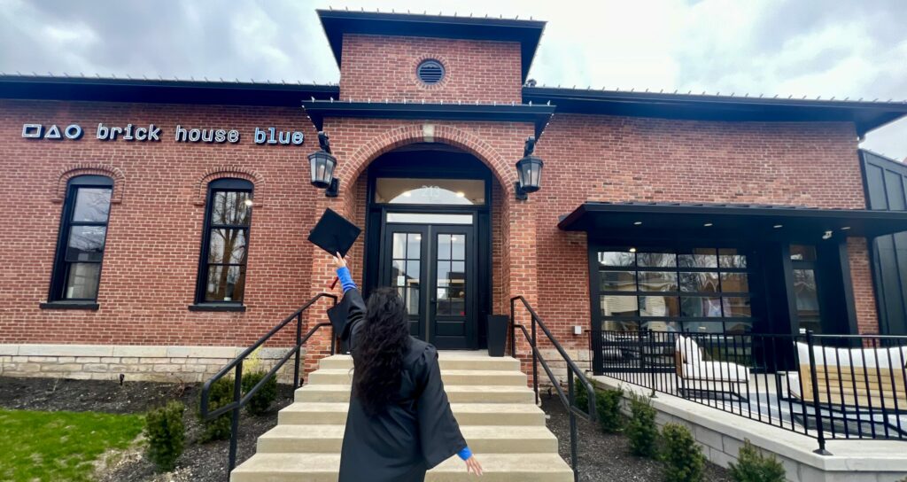 A jubilant graduate celebrating outside a charming brick building with black awnings, ideal as a graduation party venue or graduation event space