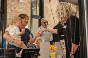 Attendees at a graduation event engaging with a staff member, with a child in arms, highlighting a family-friendly atmosphere within a venue adorned with rustic brick walls.
