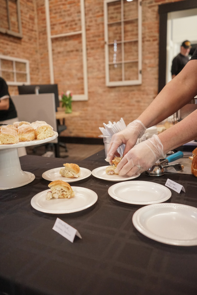 In the image, a person wearing protective gloves is serving sliced bread on white plates, arranged neatly on a table with a black tablecloth. The setting suggests a catering service at an event, with a rustic brick wall background adding charm to the venue. Small name tags in front of the bread indicate the variety or ingredients, ensuring guests can easily choose according to their preference. The environment looks inviting for attendees to enjoy a casual dining experience during a break in meetings or networking events.