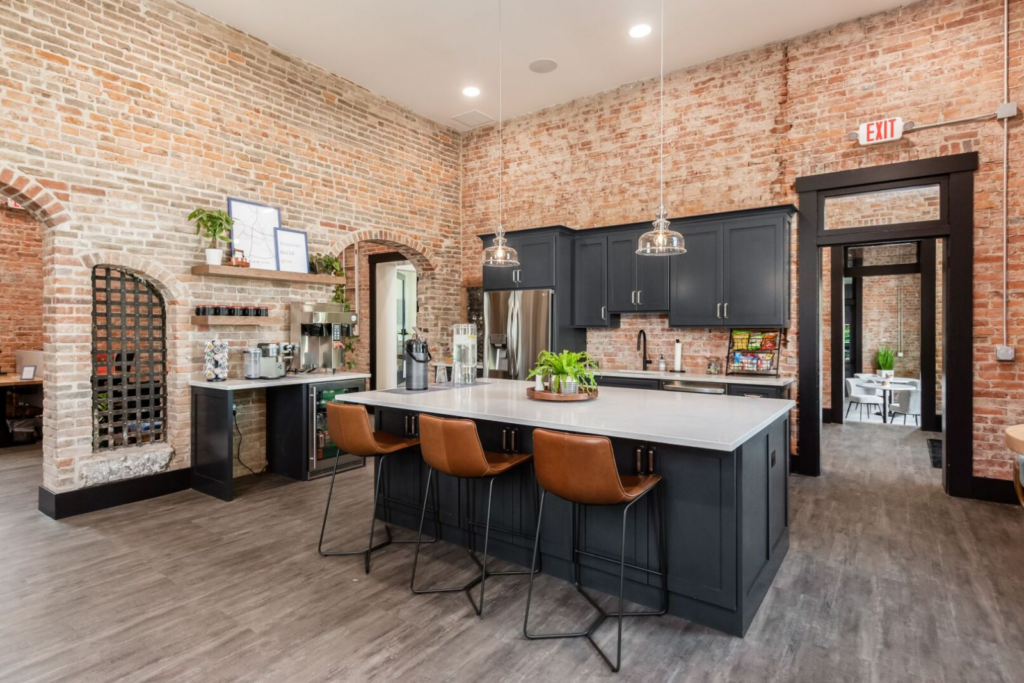 This image showcases a stylish kitchen area within a professional setting, possibly a break room or communal area in an office. It features exposed brick walls, black cabinetry, and a large central island with modern bar stools, creating a warm and inviting atmosphere. The space is designed for functionality and social interaction, complete with contemporary pendant lighting, stainless steel appliances, and neatly arranged shelves with plants and coffee-making equipment. The open archway leads to an adjoining room, suggesting an easy flow between areas in this thoughtfully designed meeting space.