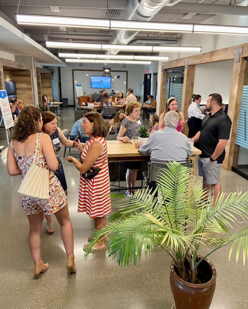 A lively gathering inside a modern meeting space, with individuals casually networking. The room is bright and airy, accented with wooden pillars, sleek furniture, and a potted palm plant adding a touch of greenery. People are spread out, some standing in conversation, others seated at tables, creating a dynamic atmosphere conducive to professional interaction and socializing.