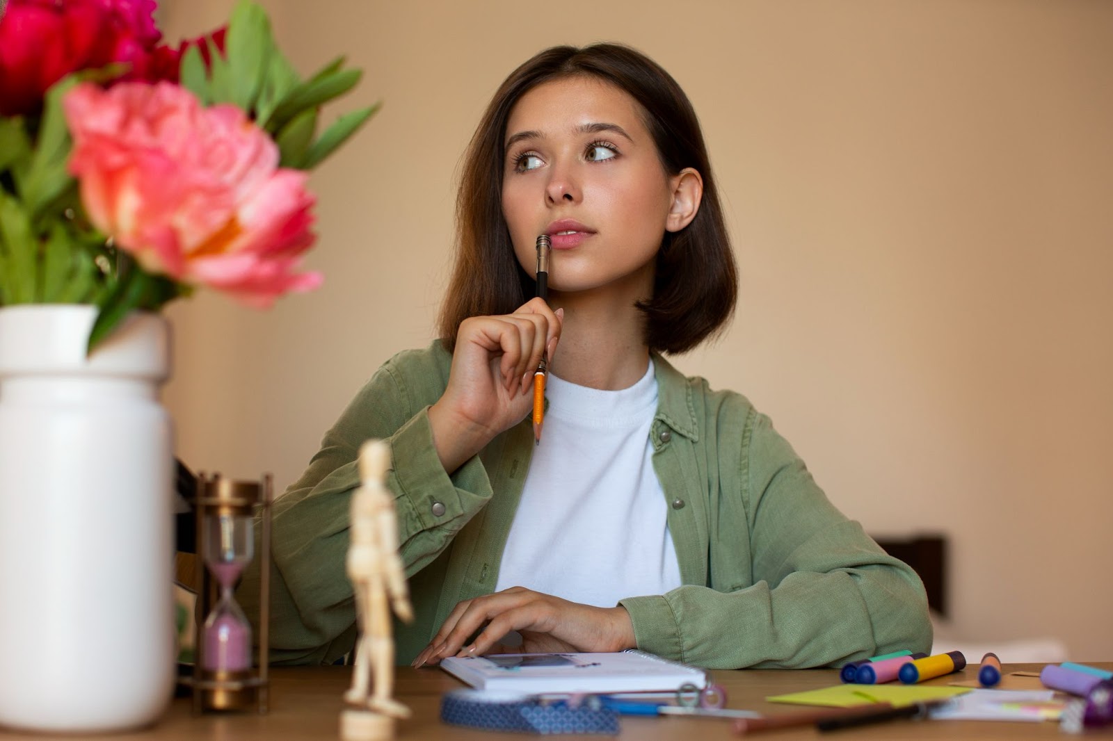 A young woman in a relaxed event planning space, deep in thought while surrounded by objects that suggest a creative brainstorming session. The prominent display of peonies and a classic hourglass on her desk add to the aesthetic of the event space, where ideas for festivities are likely being conceptualized and organized