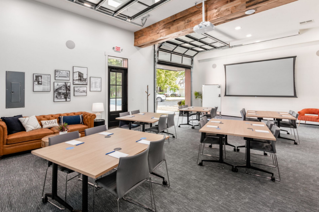 An inviting and well-lit meeting space featuring a combination of modern tables and comfortable seating arranged for collaborative work. The room is equipped with a large projection screen, suitable for presentations and workshops. Exposed wooden beams and a gallery wall of black-and-white photographs add a touch of warmth and history to the space, while the open glass door brings in natural light, creating a bright and airy atmosphere conducive to productivity and creative thinking.