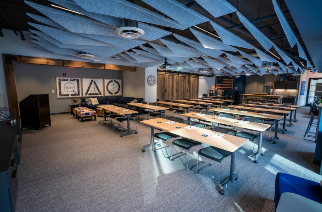 A spacious and modern meeting space with rows of tables arranged for a conference or training session. The room features contemporary design elements such as an acoustic panel ceiling, wooden accents, and a cozy lounge area with sofas. Large wall clocks and creative art pieces adorn the walls, adding character to the professional setting. The space is well-equipped for collaborative work, with a sleek, functional aesthetic and ample seating for participants.
