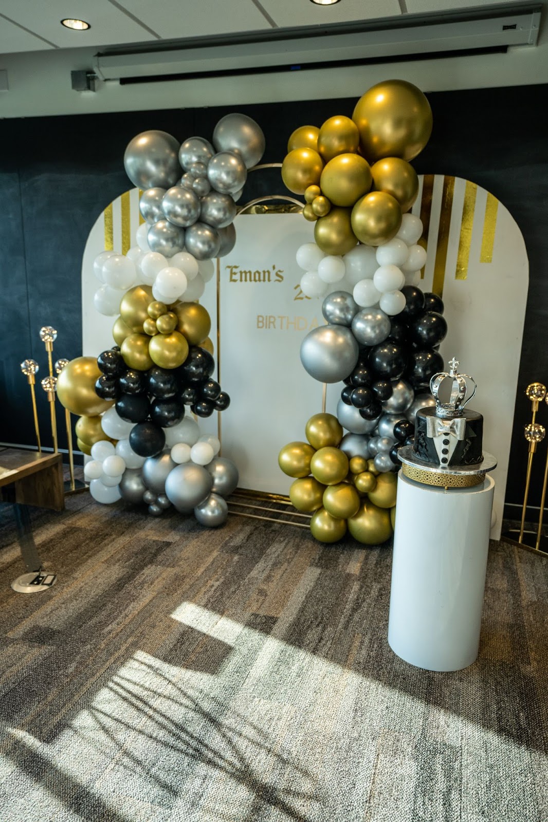 An event space decorated for a birthday celebration, with a lavish balloon arch in shades of black, gold, silver, and white balloons. The focal point is a backdrop with 'eman's birthday' printed on it, framed by the balloon arrangement. A stylish black and white birthday cake with a crown topper sits on a white pedestal in the foreground, adding a regal touch to the decor.