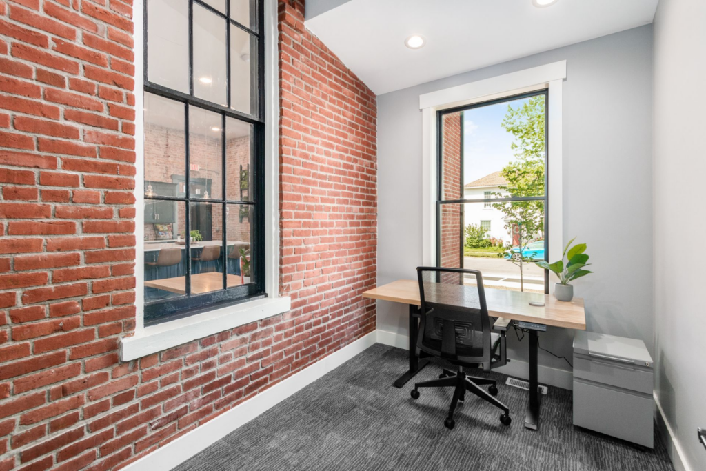 A compact office featuring a single desk and ergonomic chair inside an executive suite, with exposed brick walls and natural light from a large window