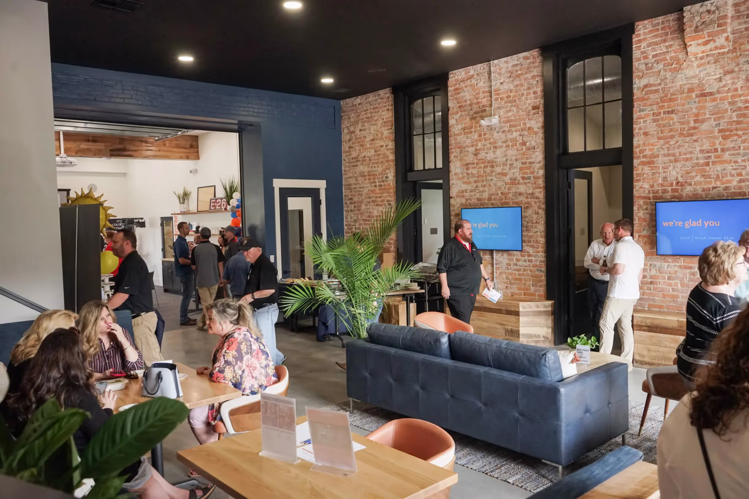 People gather and socialize in a modern, loft-style office space for rent with brick walls, sofas, plants, and tables. Some are seated and chatting, while others stand in groups. TV screens display a blue welcome message.