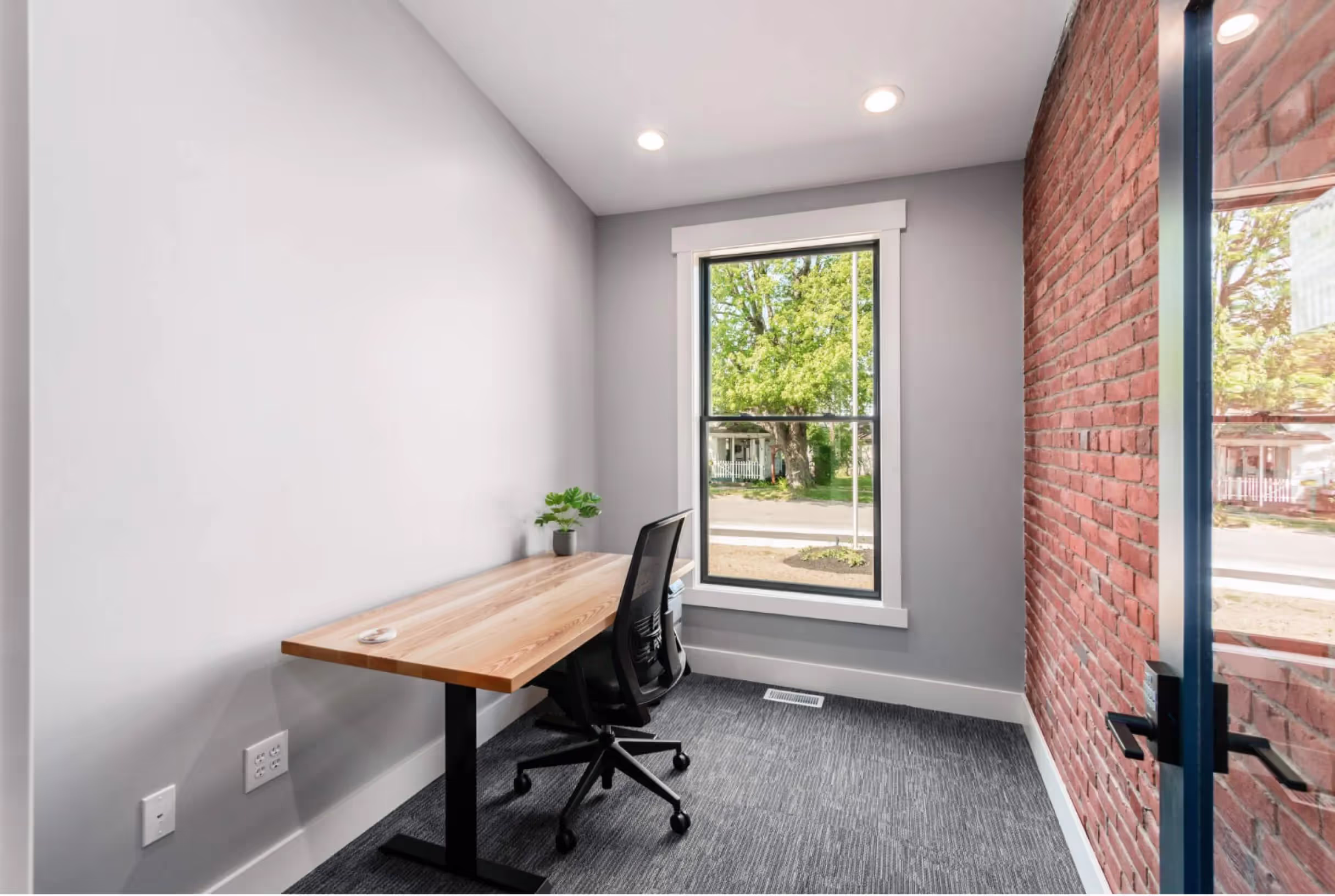 A minimalist home office with a wooden desk, black office chair, small green plant, and a large window letting in natural light; one wall is exposed red brick, the others are white—ideal inspiration for private offices or coworking space designs.