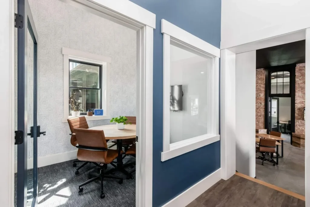 A modern coworking space with a glass-walled meeting room featuring a round table, brown chairs, and a potted plant. The hallway with blue and white walls leads to an open workspace with brick accents, ideal for collaboration.