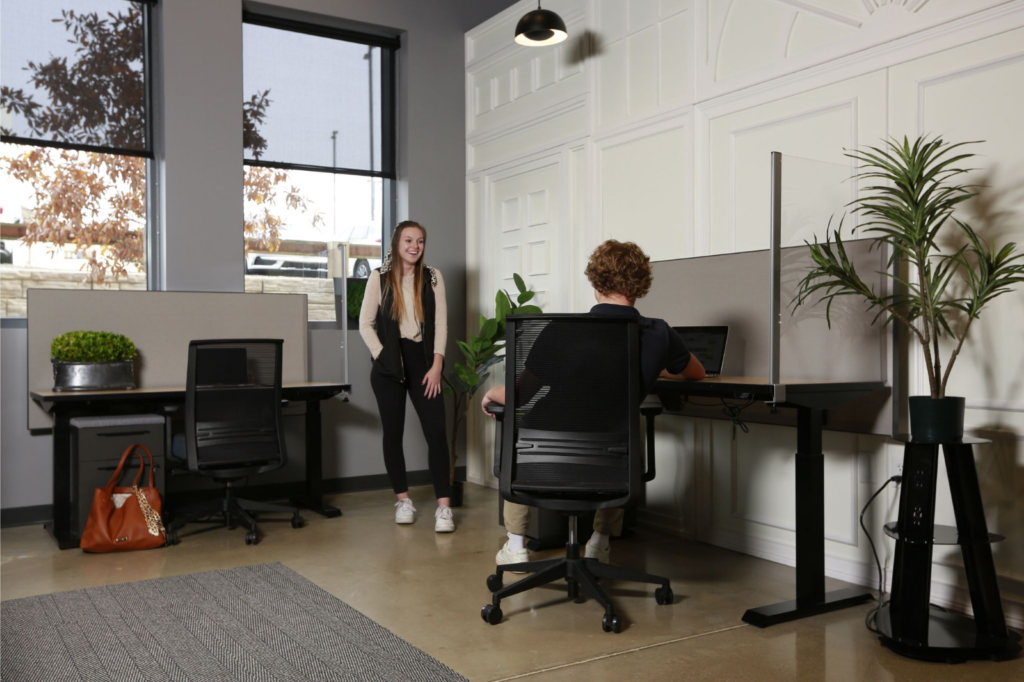 The image shows an executive suite where one person works at a desk separated by a privacy screen, while another stands nearby. The setup features ergonomic chairs, adjustable desks, and natural light through large windows. This reflects a modern approach to rent office space that combines productivity with a comfortable, welcoming atmosphere.