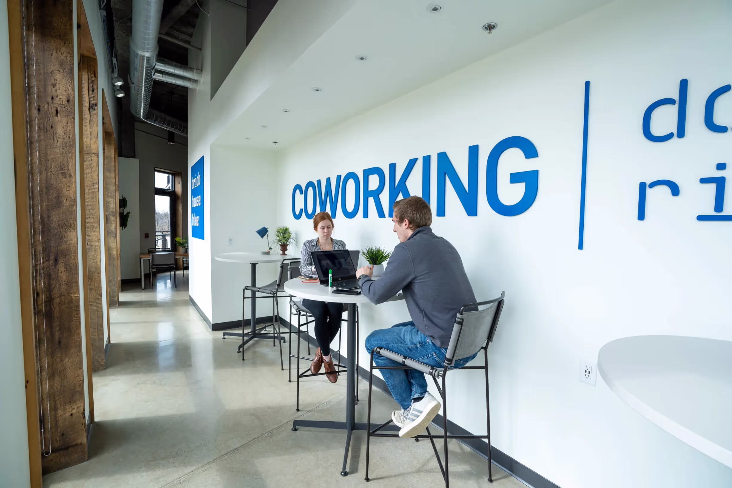 Two people sit across from each other at a high table with laptops in a modern, bright coworking space. The word COWORKING is prominently displayed on the wall behind them—ideal for collaborations or finding meeting rooms in Columbus, Ohio.