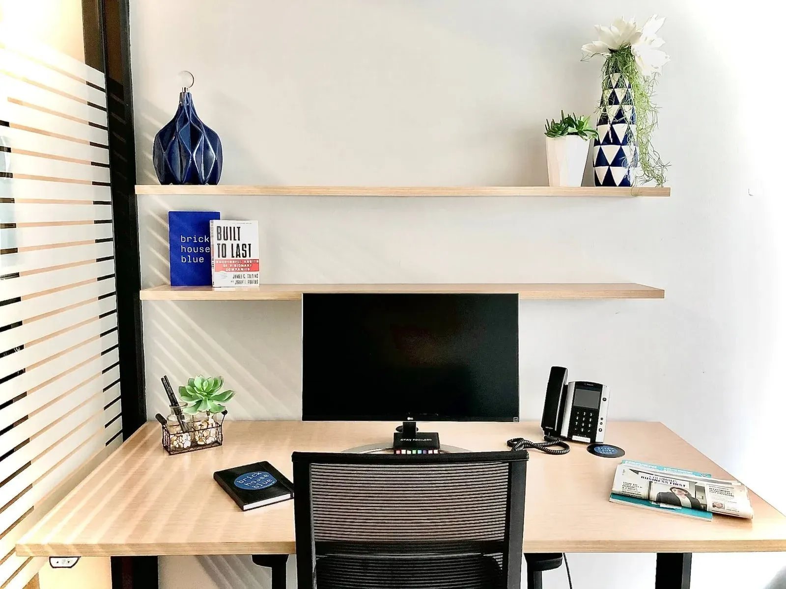 A modern, minimalist desk setup perfect for a coworking space, featuring a computer monitor, phone, notepad, and plants. Two wooden shelves above hold books, vases, and flowers. A comfortable chair completes this stylish workspace.