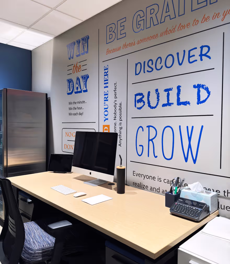 A modern office workspace with a computer, keyboard, pen holder, tissue box, and calculator on a desk—ideal for businesses seeking a private office. The wall displays motivational phrases like “WIN THE DAY,” “DISCOVER,” “BUILD,” and “GROW.”.