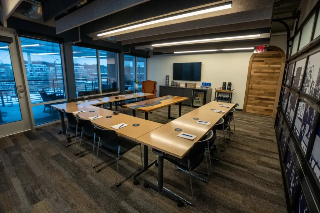 Modern conference room with a U-shaped table setup, black chairs, notepads and pens on the tables, a large screen, bookshelf, orange chair, and large windows with a city view—ideal meeting rooms or event spaces in Columbus Ohio.