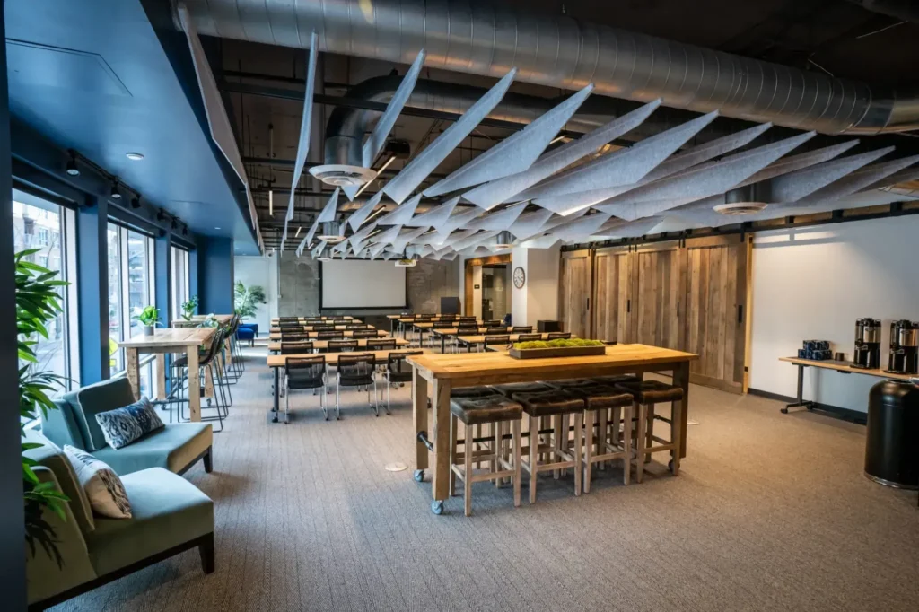 Modern, industrial-style office space for rent featuring meeting rooms with rows of tables and chairs, a central high table with stools, wooden sliding doors, coffee station, sofa, large windows, and geometric ceiling panels for sound absorption.