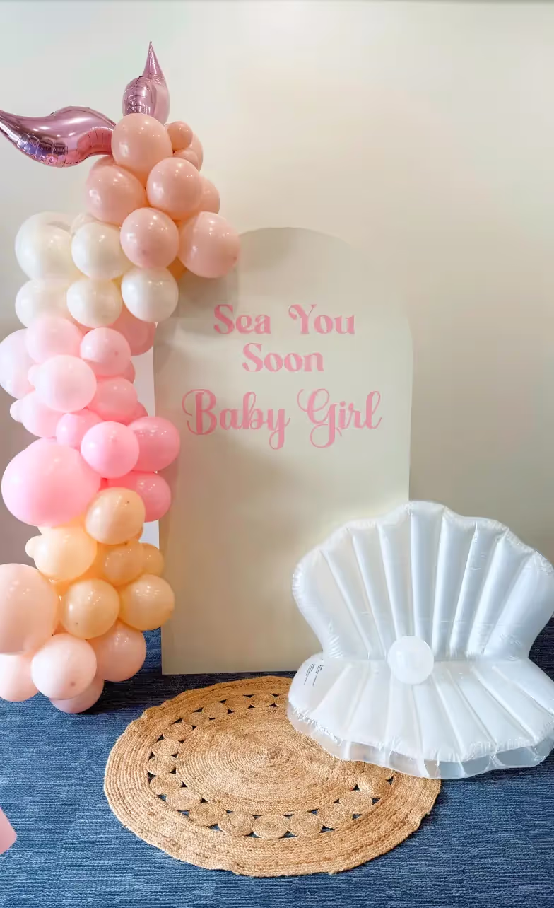A baby shower setup in one of the charming event spaces Columbus Ohio offers, with pink and peach balloons shaped like a mermaid tail, a “Sea You Soon Baby Girl” sign, a round woven rug, and an inflatable white seashell chair on blue carpet.