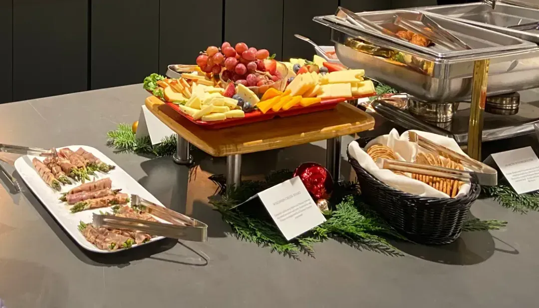 A buffet table in a coworking space, featuring assorted cheeses, fruits, crackers, and sliced meats in chafing dishes, beautifully decorated with greenery and holiday ornaments—perfect for gatherings or meetings.