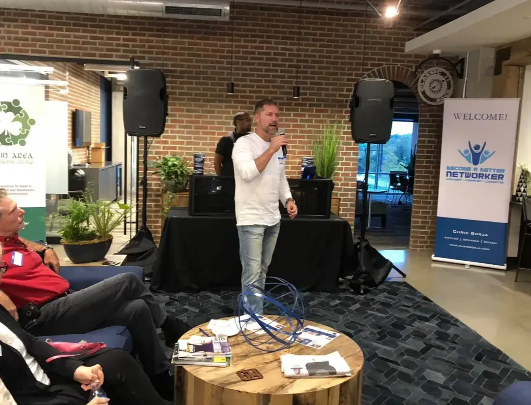 A man in a white shirt and jeans speaks into a microphone in a modern room with brick walls, ideal for private offices. People are seated, and banners and plants decorate the space. Audio equipment is set up behind him.