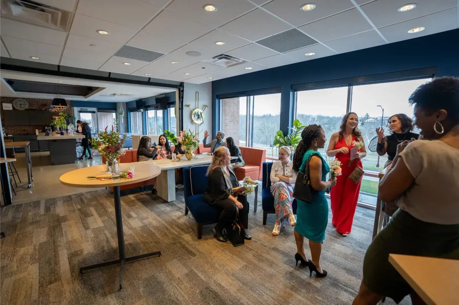 A group of people socialize in a modern, well-lit lounge with large windows, tables, and flowers—an inviting office space for rent where some are seated, others stand and chat, creating a lively and welcoming atmosphere.