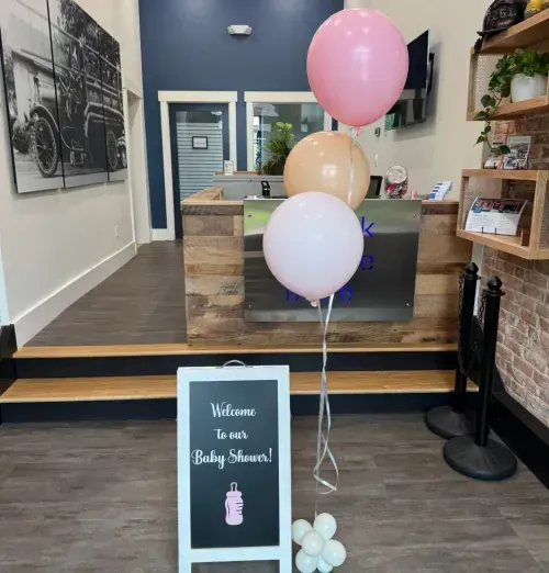 A small sign with Welcome to our Baby Shower! stands in front of a reception desk, beside four pastel balloons and a small white balloon cluster. The room, perfect for event spaces Columbus Ohio, has wood accents and black-and-white wall art.