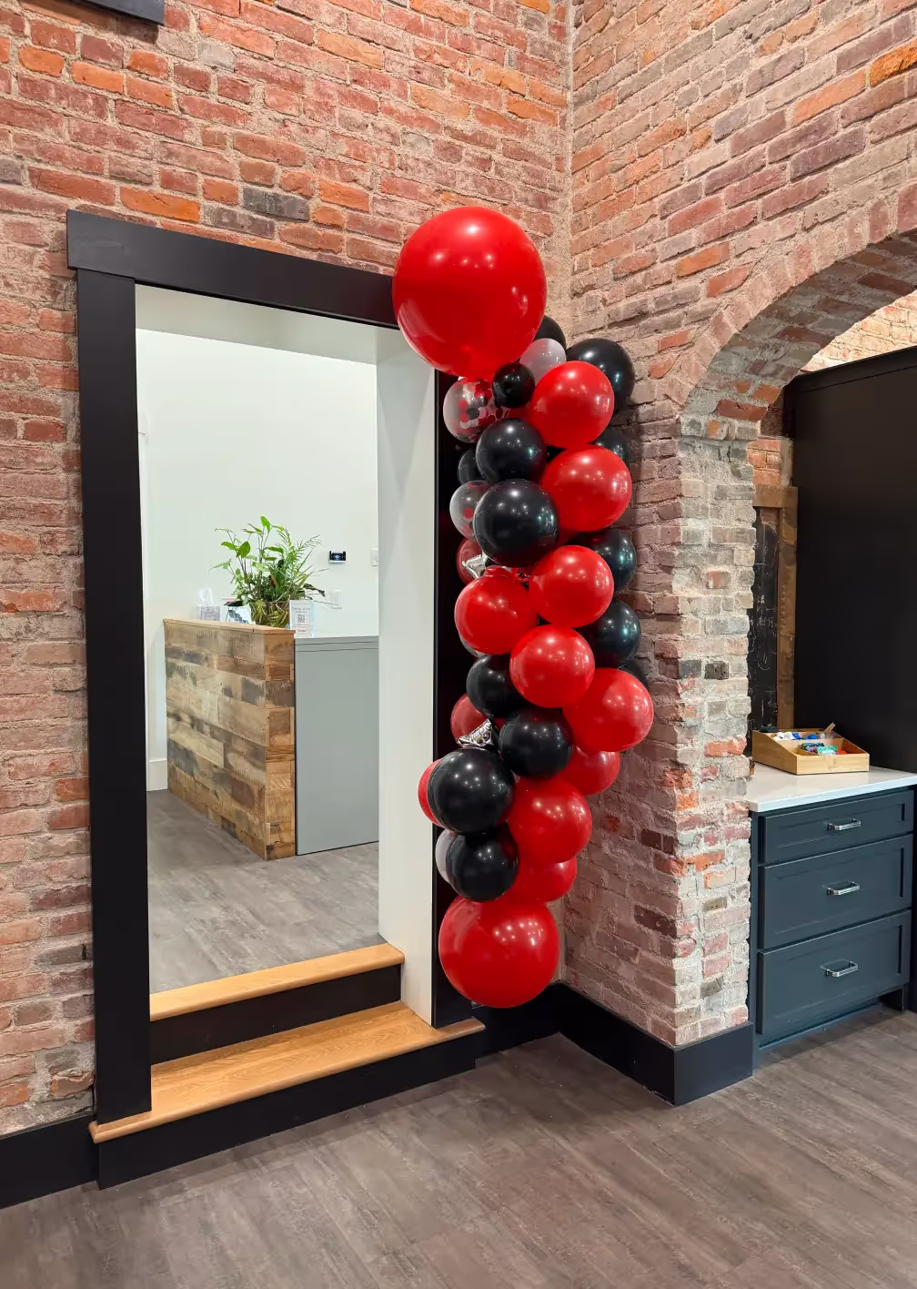 A bunch of red and black balloons is attached to the corner of a doorway in a brick-walled coworking space with wood flooring. A plant sits on a rustic wooden reception desk in the background.