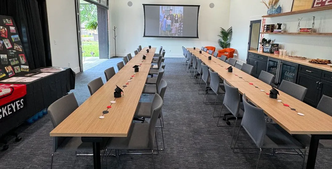 A conference room in a coworking space features two long tables arranged in a U-shape, each set with black napkins and circular tokens. A projector displays an image, with chairs, decorations, and refreshments around the room.