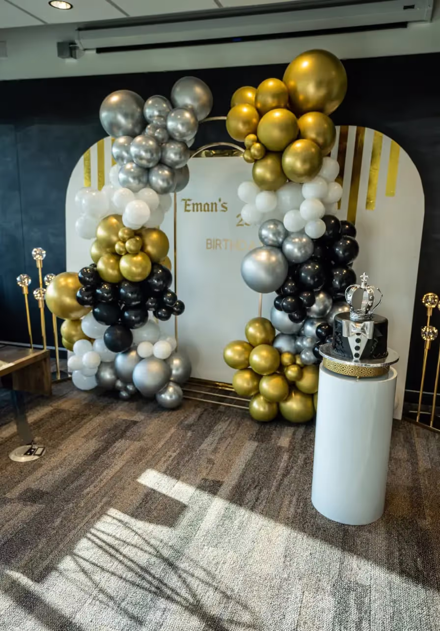 A birthday party setup in one of the finest event spaces Columbus Ohio offers, with a gold, silver, black, and white balloon arch, a white panel reading Emans Birthday, and a black cake with crown topper on a stylish white pedestal.