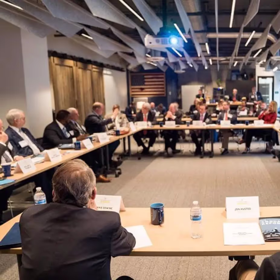 People in business attire sit around a large U-shaped conference table in a modern meeting room, engaged in discussion. Documents, cups, and water bottles are on the tables, showcasing the versatile event spaces Columbus Ohio has to offer. An American flag hangs on the back wall.