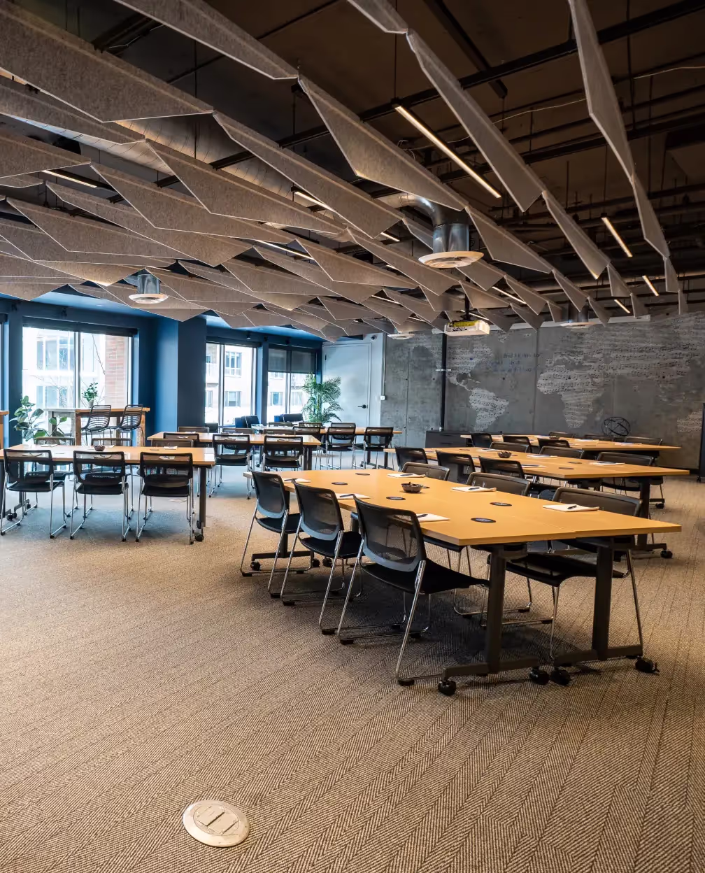 A modern coworking space featuring several empty tables and chairs on wheels, large windows streaming natural light, and geometric acoustic panels hanging from the ceiling—perfect for meeting rooms or event spaces in Columbus, Ohio.