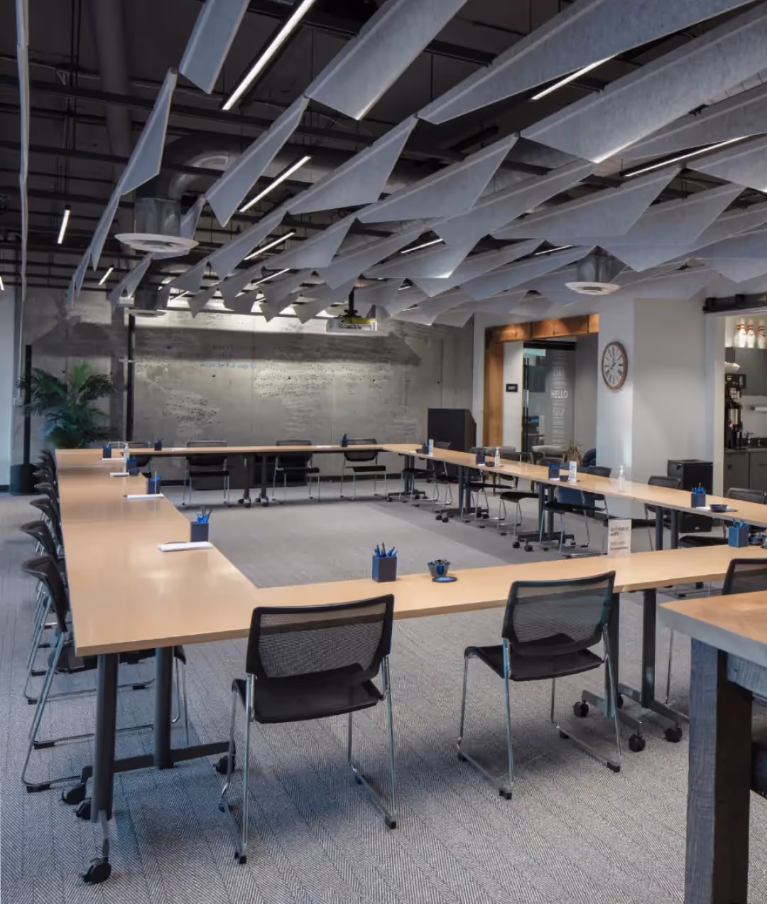 Modern office meeting room with U-shaped wooden tables, black mesh chairs, ceiling acoustic panels, and various office supplies. Ideal for teams seeking office space for rent, the well-lit room features industrial decor and a visible whiteboard wall.