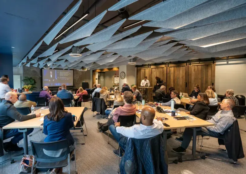 A group of people sit at tables in a modern conference room with wood accents and gray ceiling panels, using meeting rooms designed for collaboration as they watch a presentation on a screen in this casual, professional atmosphere.