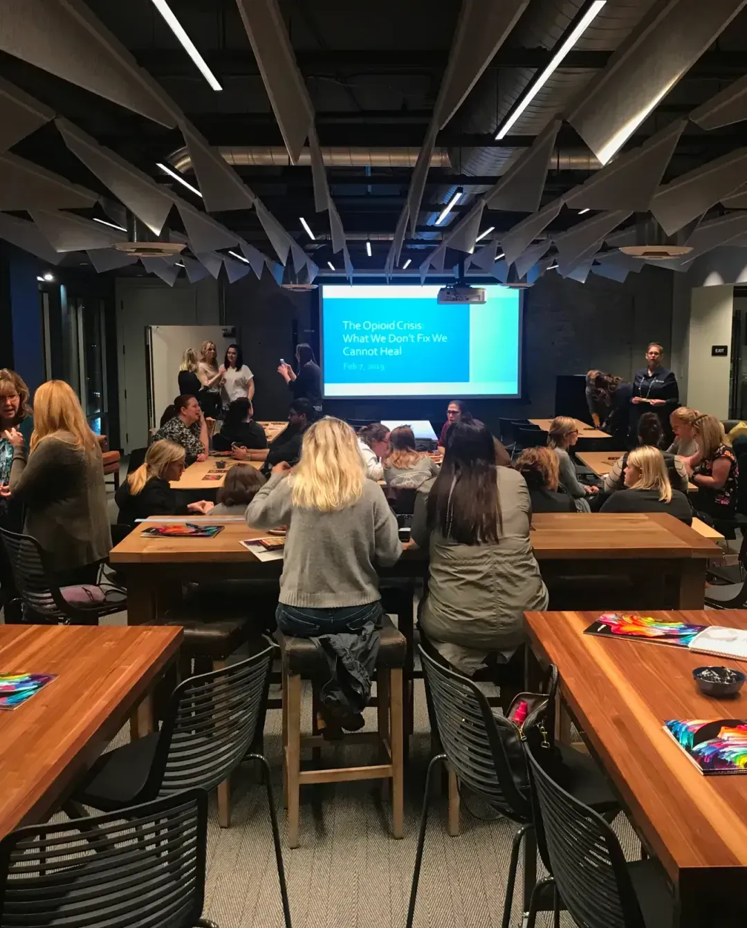 A group of people sit and stand around tables in a modern coworking space meeting room, facing a large screen displaying a blue presentation slide titled “The Opioid Crisis: What We Don’t Fix We Cannot Heal.”.
