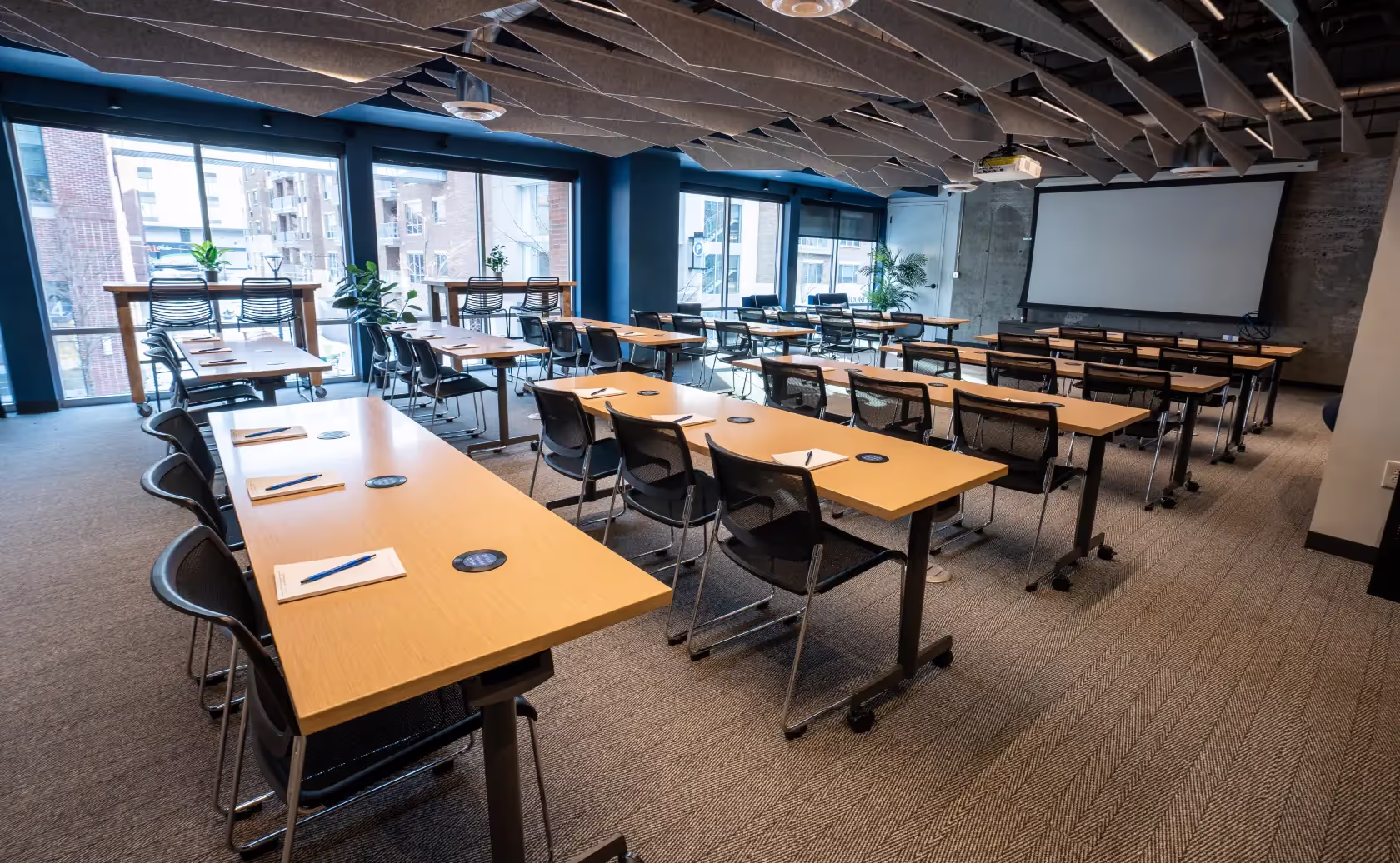 Modern conference room in a coworking space with rows of tables and chairs, notepads and pens on each desk, large windows, plants, and a projection screen. Ceiling has geometric sound panels. This event space in Columbus, Ohio is brightly lit and empty.