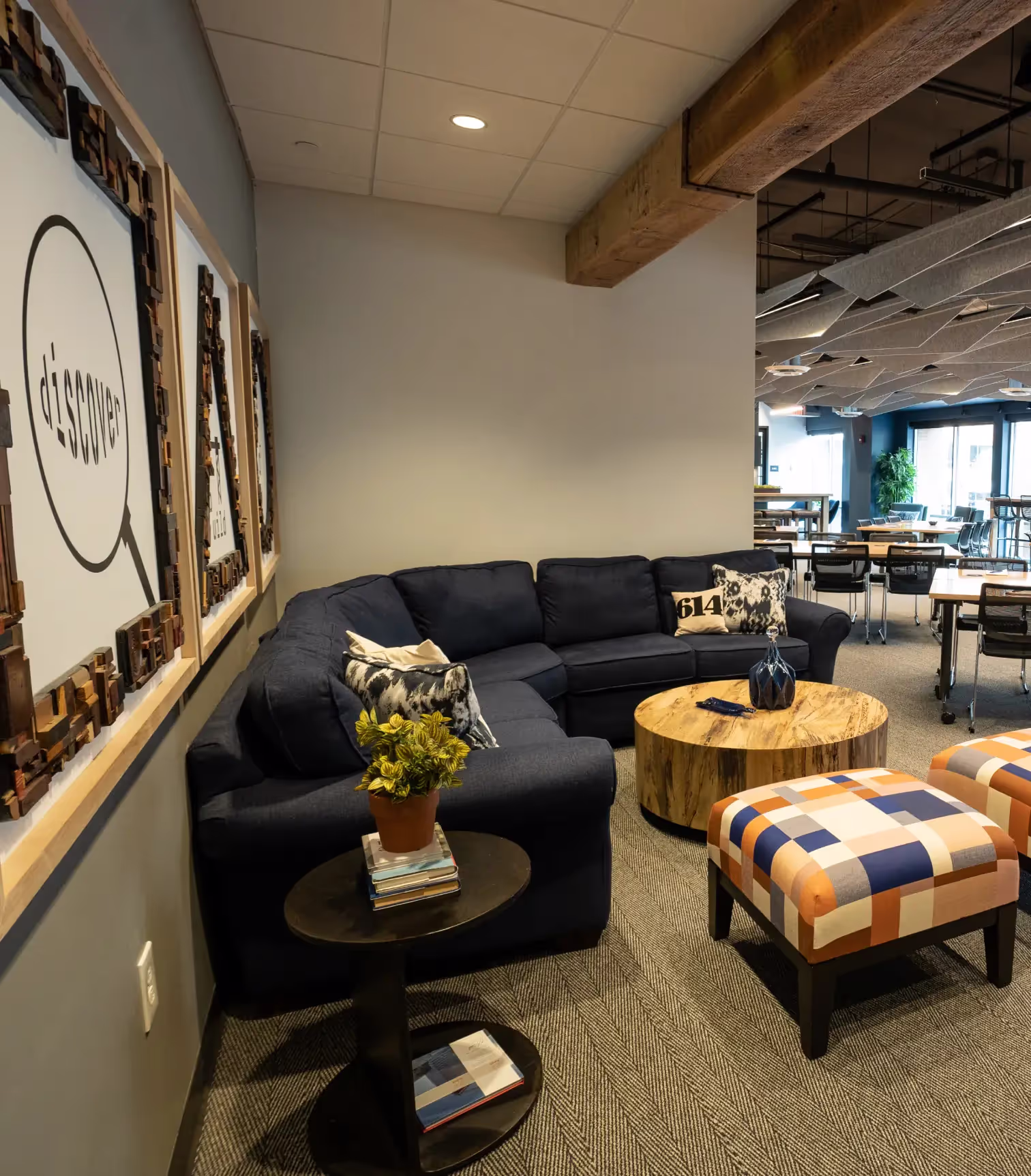 A cozy office lounge in a coworking space with a navy sectional sofa, plaid ottomans, a round wooden coffee table, and framed wall art. A side table with a plant and books sits nearby, with tables and chairs and meeting rooms visible in the background.