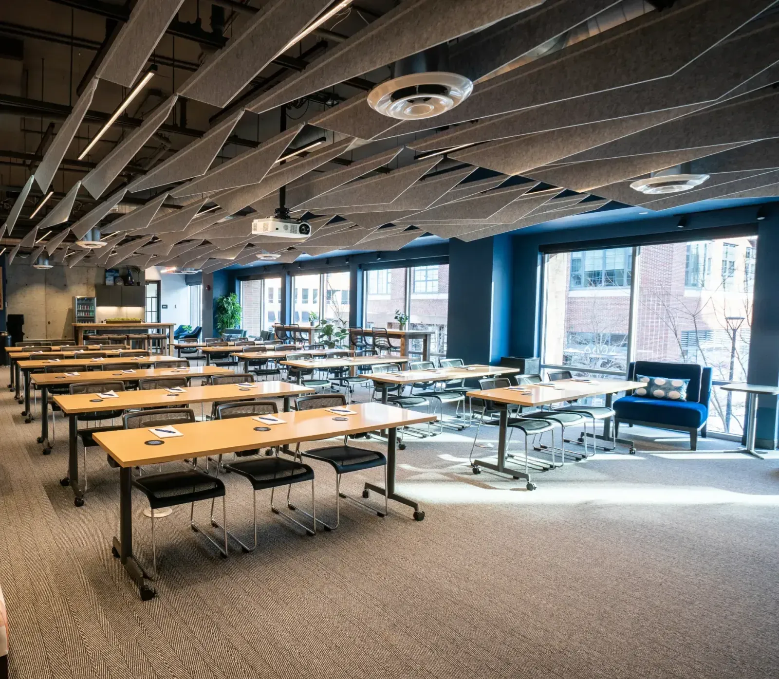 Spacious, modern coworking space or classroom with long tables, black chairs, large windows letting in natural light, ceiling acoustic panels, plus a lounge area featuring a blue sofa and cushions. Ideal for meeting rooms or event spaces in Columbus, Ohio.