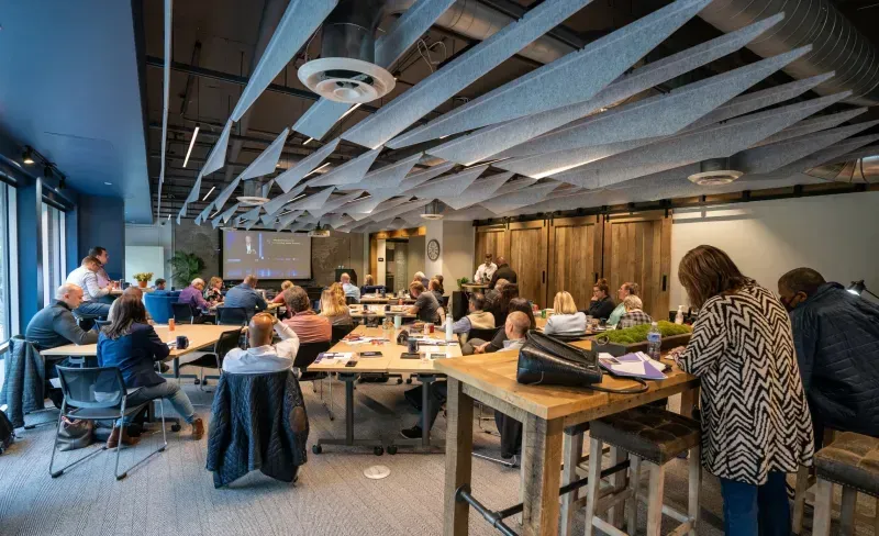 A group of people sit at tables and listen to a presentation in a modern coworking space with exposed ceilings and wooden accents. A presenter stands near a screen displaying slides at the front of the room.
