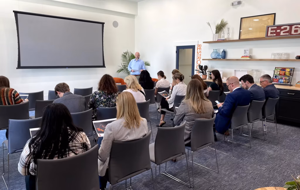 A man stands at the front of a modern conference room—ideal for event spaces Columbus Ohio—speaking to an audience seated in rows of gray chairs. A large projector screen and decorative shelves are in the background.