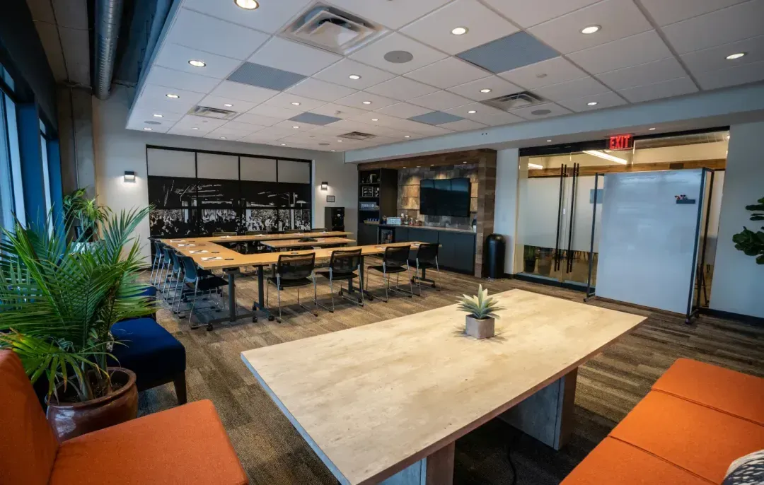 Modern conference room with long tables and chairs, a kitchenette, whiteboard, and potted plants. This bright office space for rent features large windows, carpeted floors, and overhead lighting—ideal for meetings or as a virtual business address.