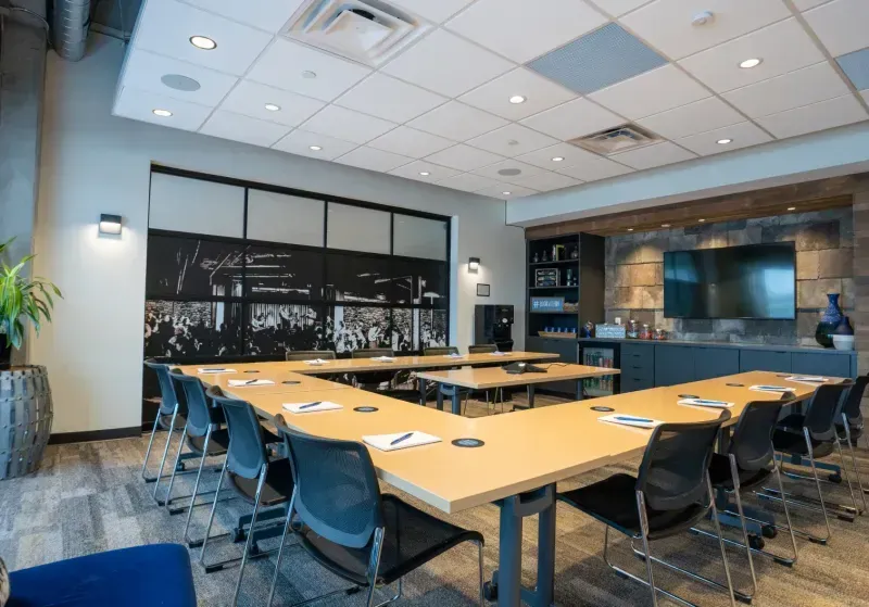 Modern conference room with U-shaped tables, black chairs, notepads and pens, a wall-mounted TV, shelves, a plant, and a large black-and-white mural. This well-lit office space for rent is organized and ideal for meetings in Columbus, Ohio.