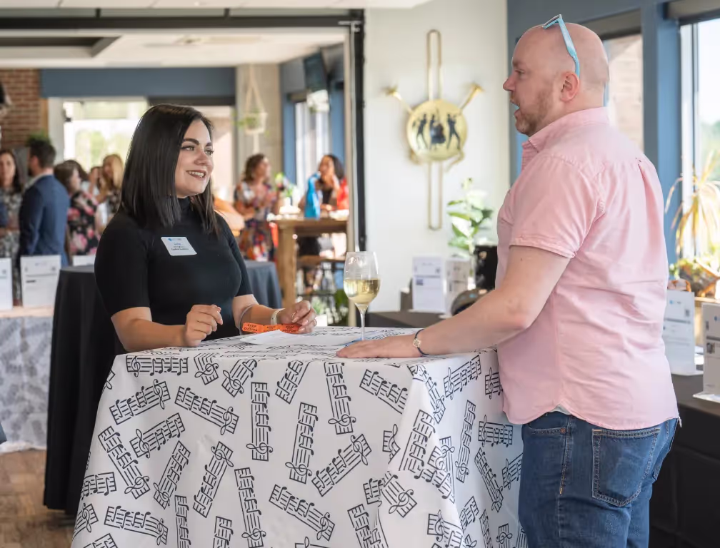 A woman and a man stand at a tall table covered with a patterned cloth, chatting at a social event in one of the top event spaces Columbus Ohio offers. The woman is smiling, and there’s a glass of white wine on the table as others mingle in the background.