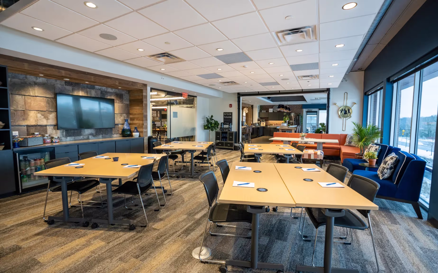 A modern office meeting room with several tables and chairs in clusters, each set with paper and pens. Large windows let in natural light, and there are plants, a TV screen, lounge seating, and private offices in the background.
