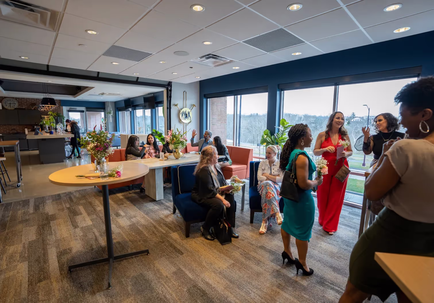 A group of people in formal attire are mingling and chatting in a spacious, modern lounge with large windows, flowers on tables, and natural light filling one of the finest event spaces Columbus Ohio has to offer.