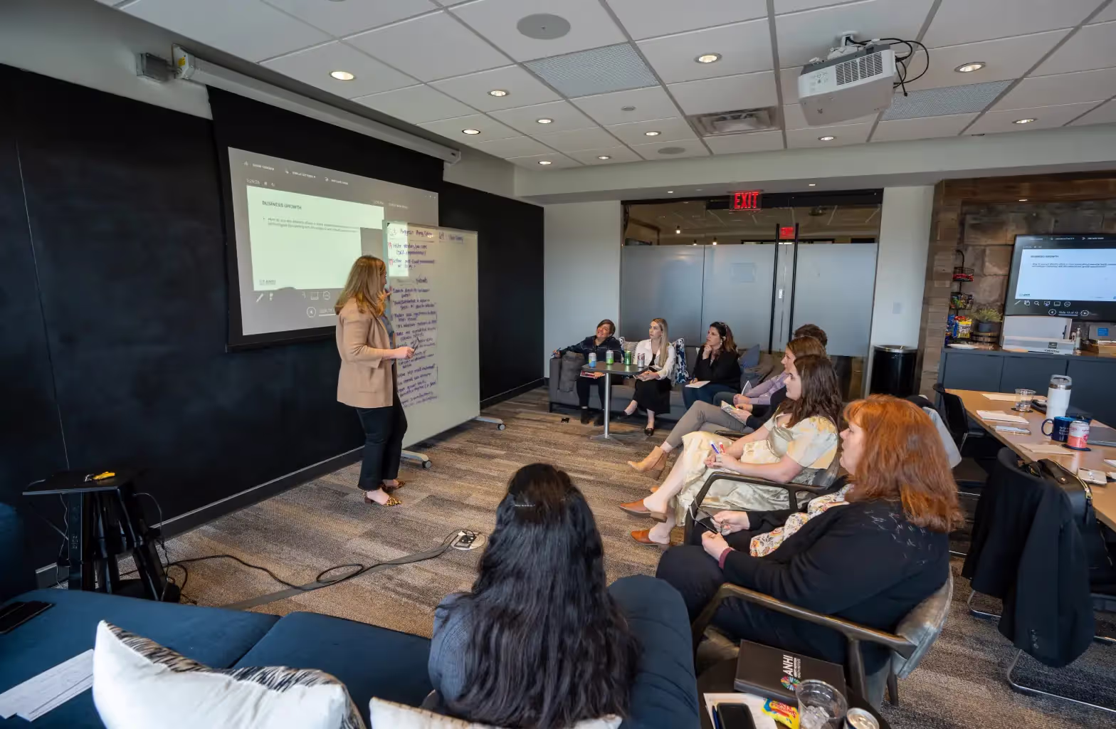 A woman stands near a screen and whiteboard, presenting to a group of seated people in a modern conference room—one of several meeting rooms available in this contemporary office space for rent. Attendees listen attentively, with laptops and notebooks visible.