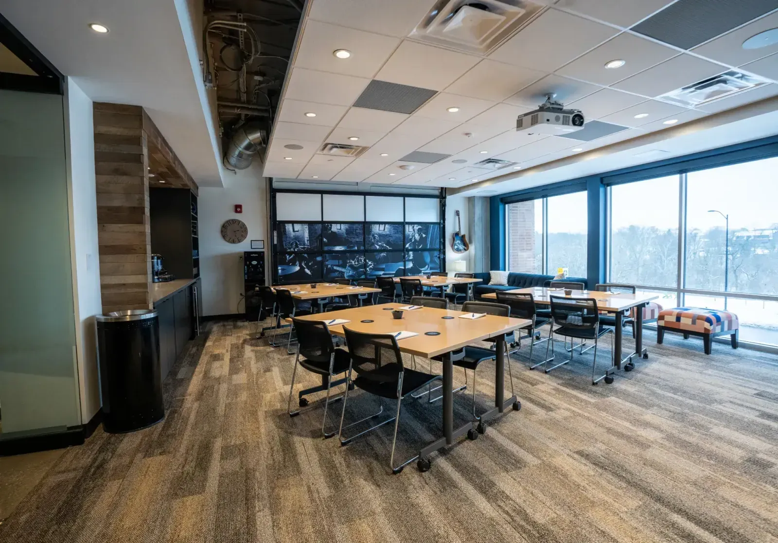 A modern coworking space with large windows, group tables and chairs, ceiling projector, and wall-mounted monitors. Natural light fills this open, inviting office space for rent—perfect for collaboration and productivity.