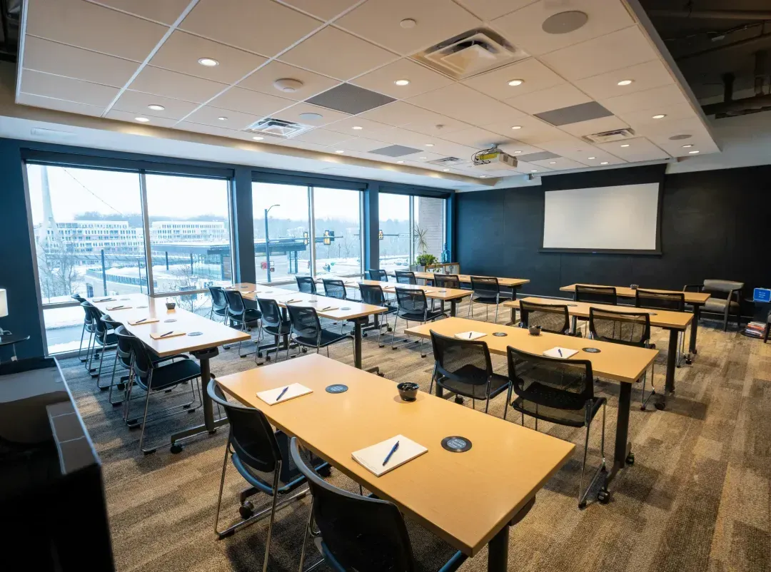 A modern, well-lit classroom with large windows, several rows of desks and chairs, notebooks and pens on each desk—ideal for groups seeking flexible event spaces in Columbus, Ohio—with a projector and a projection screen at the front of the room.