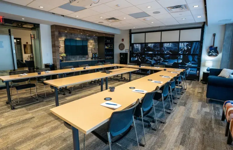 A modern conference room with U-shaped tables, black chairs, and notepads with pens at each seat—perfectly situated within a coworking space. The room features large windows, a TV, wall art, and a guitar hanging on the wall.