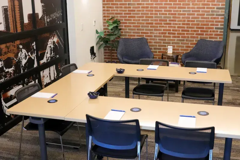 A small meeting room with U-shaped tables, black chairs, notepads, and pens—ideal for those seeking meeting rooms or event spaces in Columbus, Ohio. Two blue armchairs, a plant, and black and white wall art add a welcoming touch.