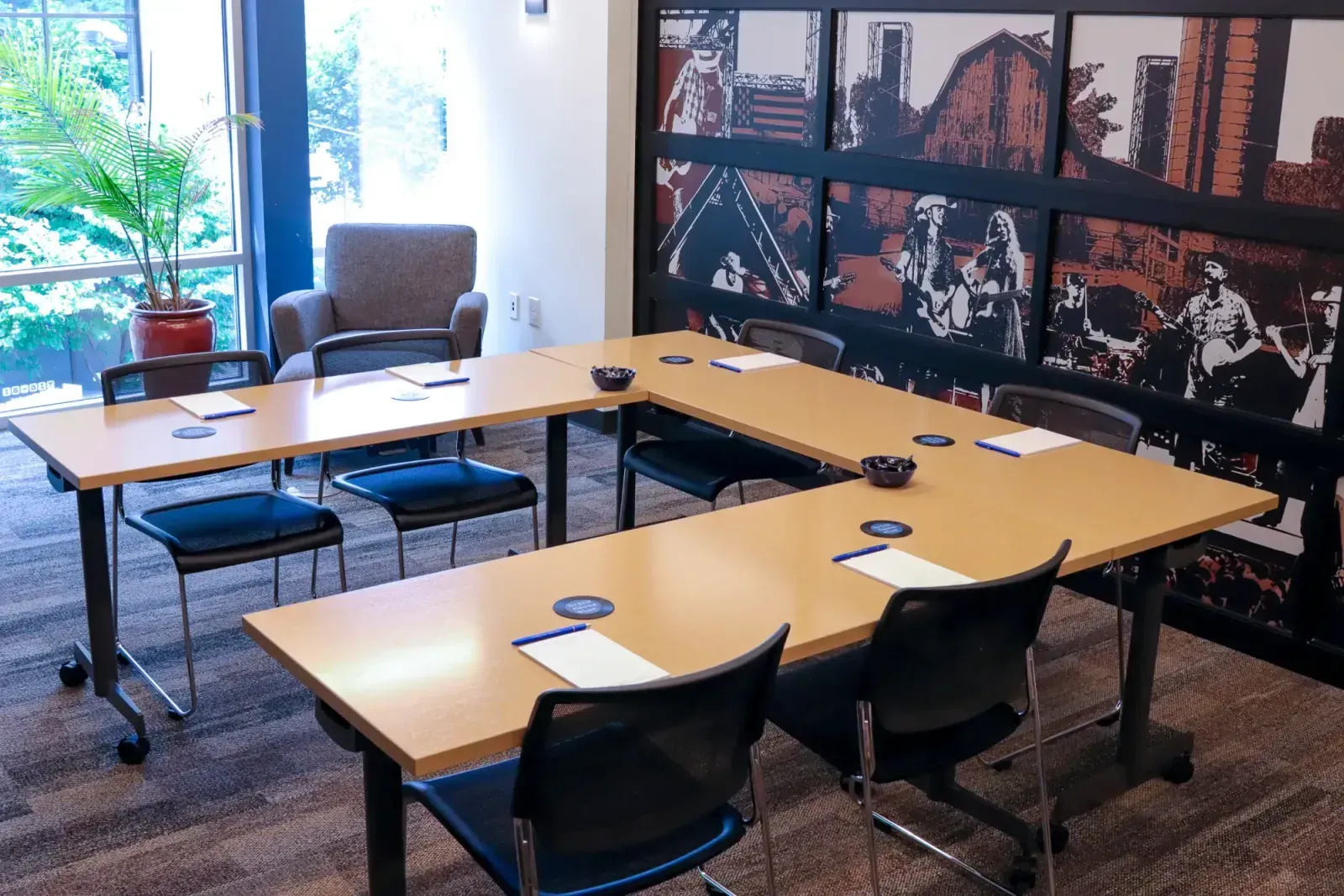 A conference room with U-shaped tables, black chairs, notepads, and coasters—perfect for meeting rooms or event spaces in Columbus, Ohio. A potted plant and armchair sit by large windows, with a vibrant mural of musicians on the back wall.