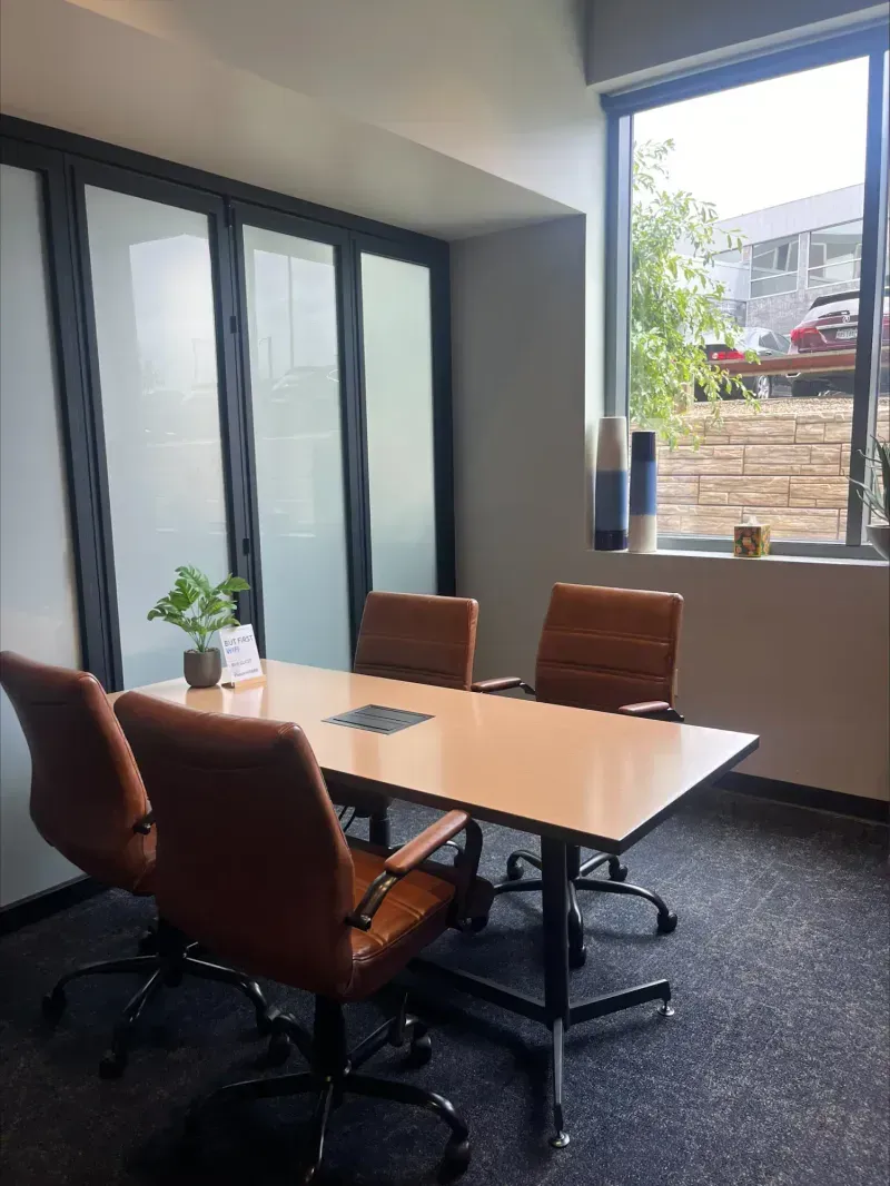 A modern coworking space meeting room with four brown chairs around a rectangular table, a small potted plant, a sign, frosted glass divider panels, and a large window letting in natural light.